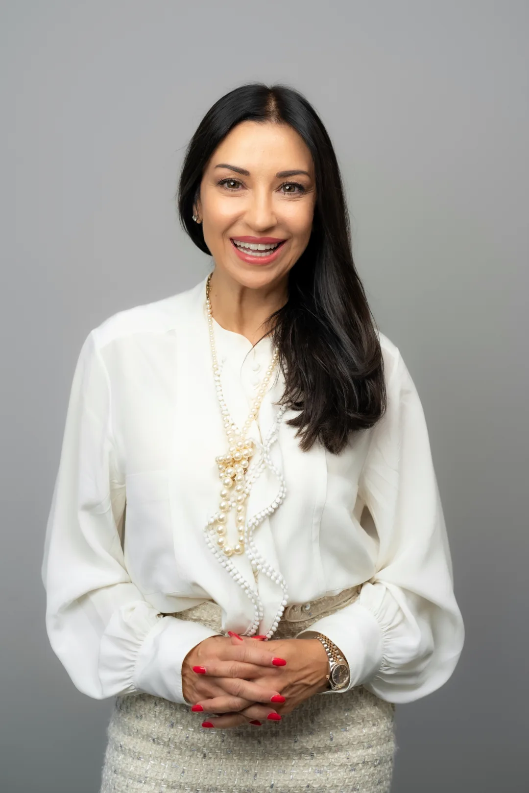 Pavlina Petrova, founder of ZaraConsult, smiling to camera in a cream silk blouse and two strands of pearls, hands clasped, against a plain grey studio backdrop